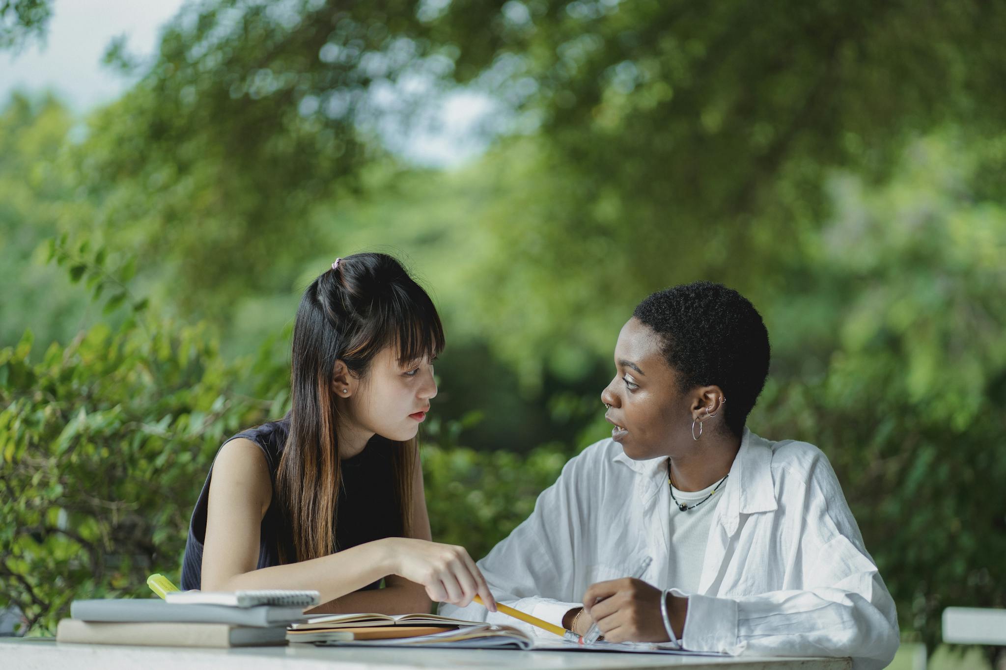 Focused multiracial female students in casual clothes sitting at table with textbooks and copybooks and discussing home assignment together while studying in lush summer garden